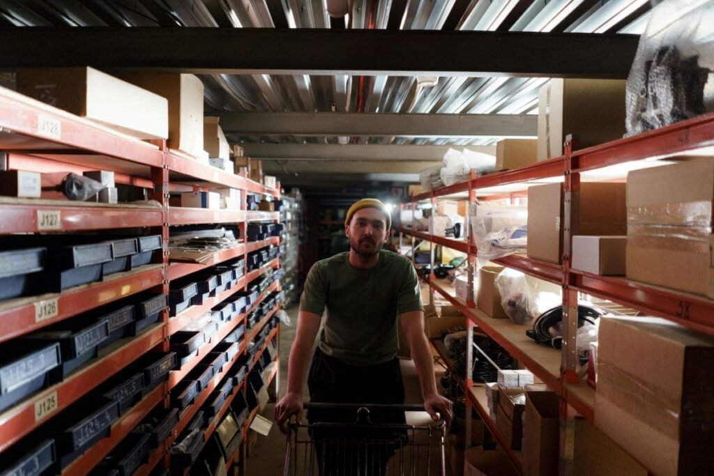 Warehouse worker pushing a cart between shelves stocked with small bins and boxes