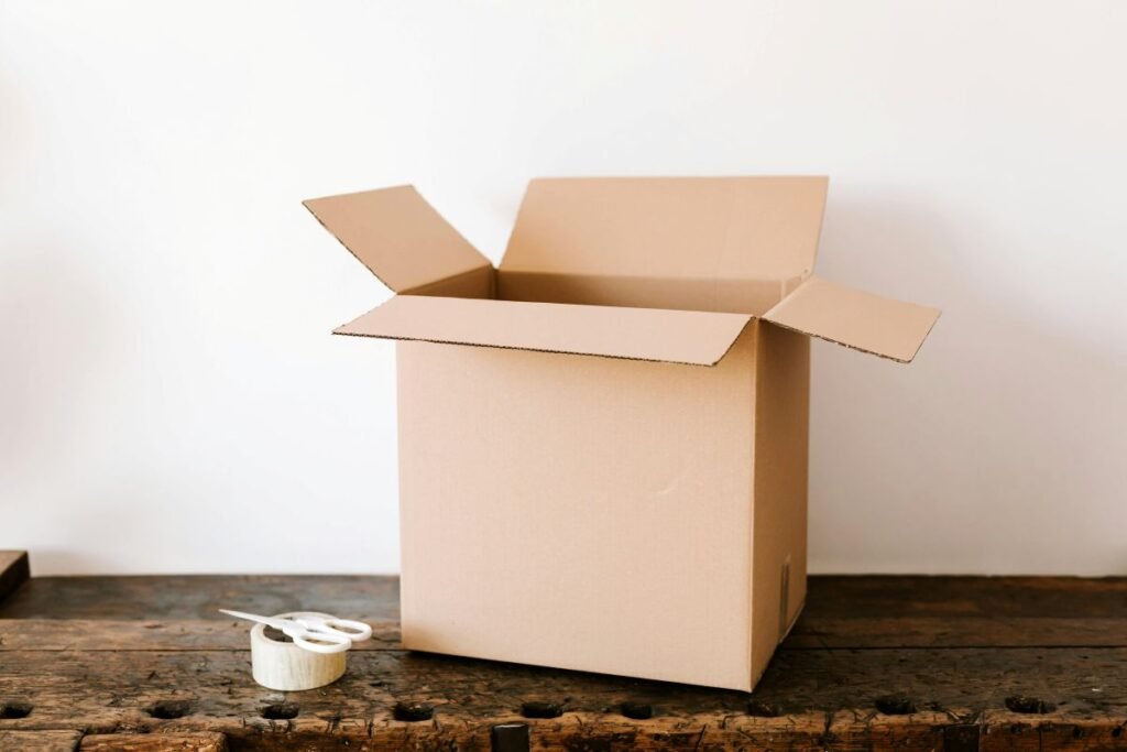 Open cardboard box on a wooden workbench with scissors and tape, demonstrating packaging preparation and the use of recycled cardboard for shipping.