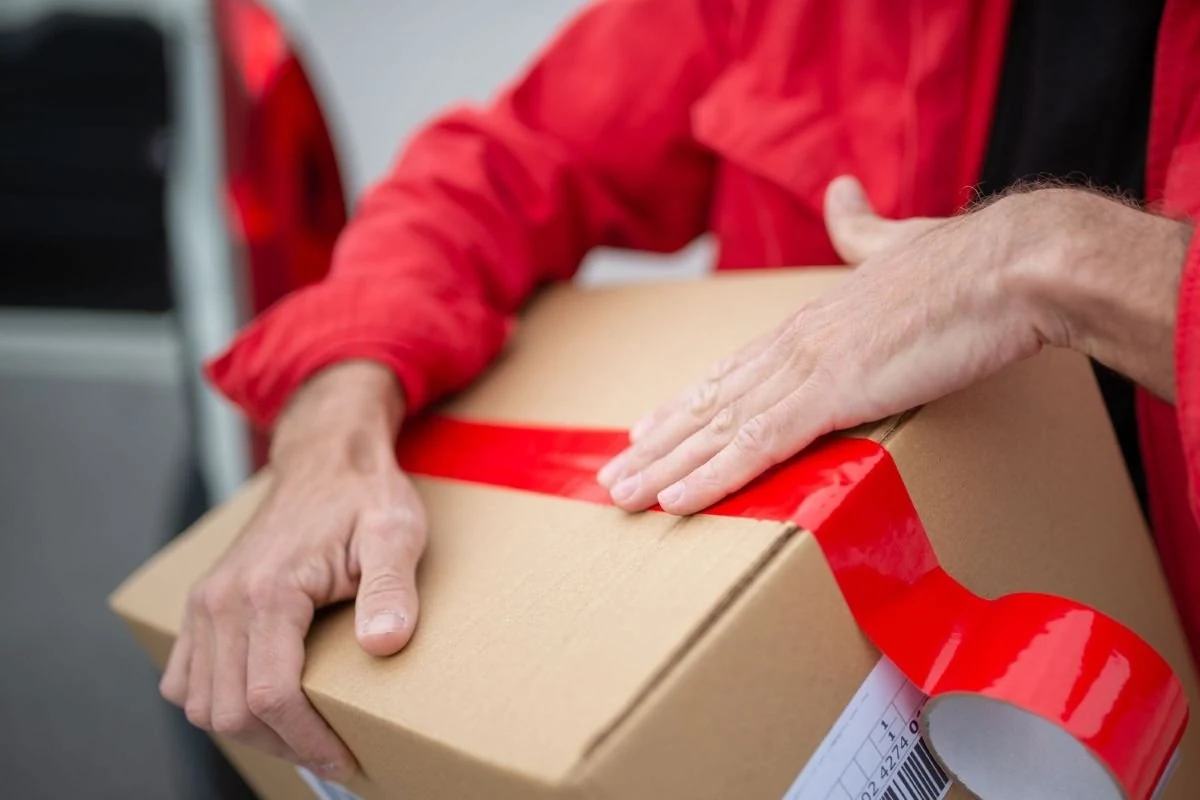 Person pressing red packing tape along the top seam of a cardboard box.