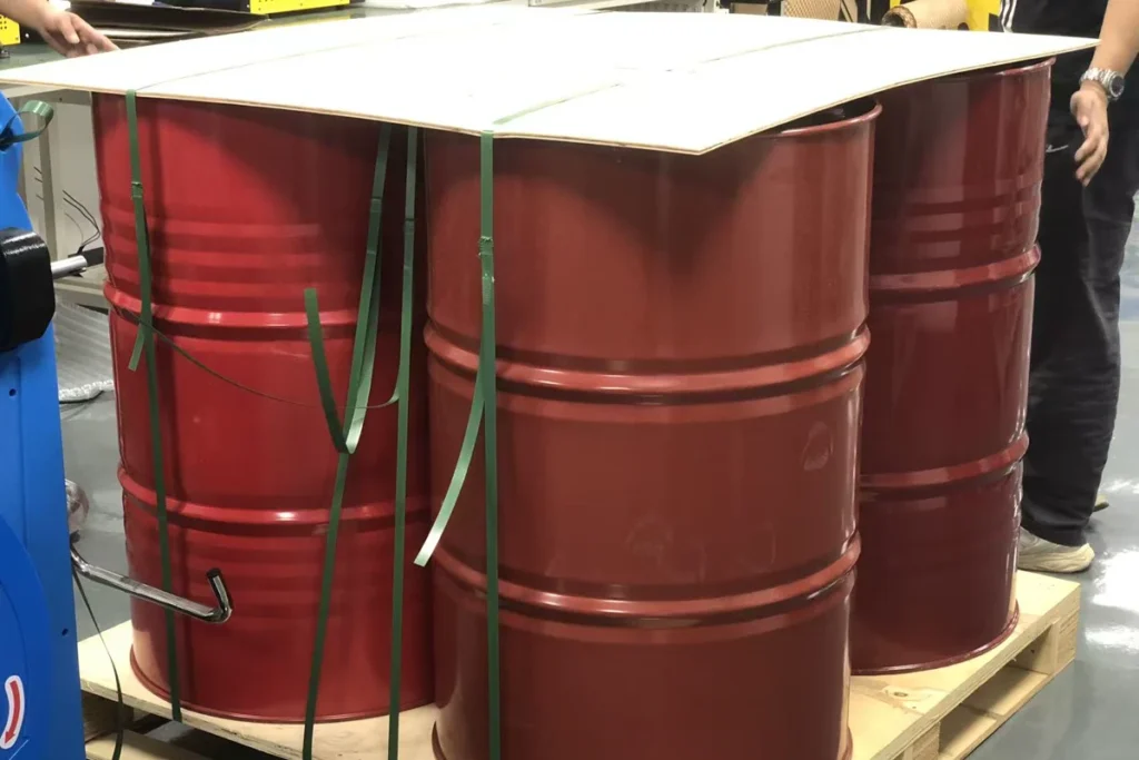 Green straps pass over a flat board spanning red steel drums on a wooden pallet.