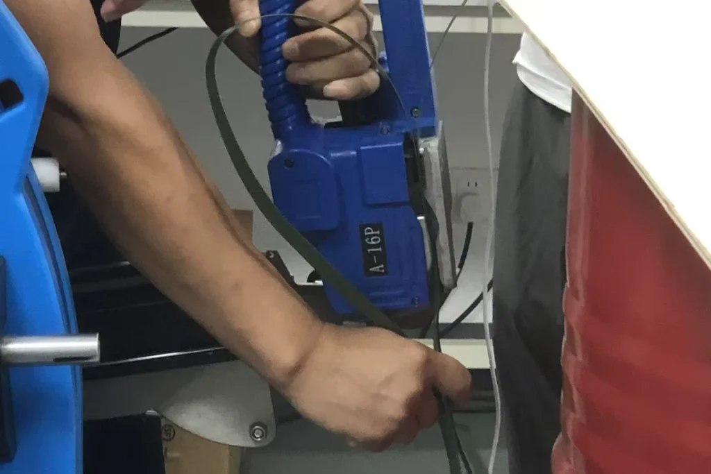 Operator handling a blue strapping tool and green strap beside a red steel drum.