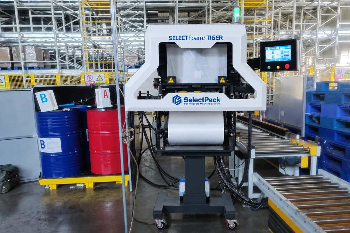 SelectPack TIGER Gen 2 foam packaging machine beside A/B chemical drums and roller conveyors in a factory setting.