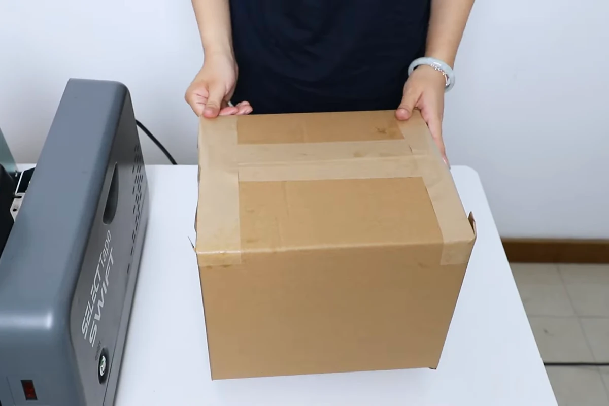 Manos que sostienen una caja sellada con cinta adhesiva marrón junto a un dispensador gris Swift.